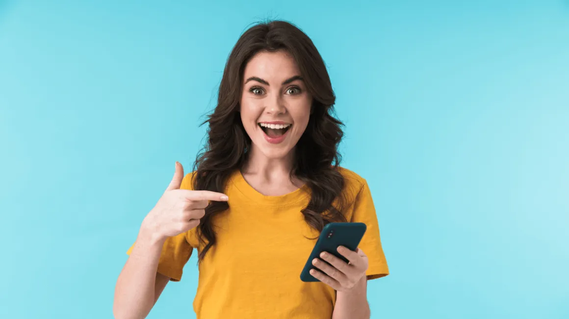 Zadowolona kobieta w żółtej koszulce patrzy w telefon na niebieskim tle